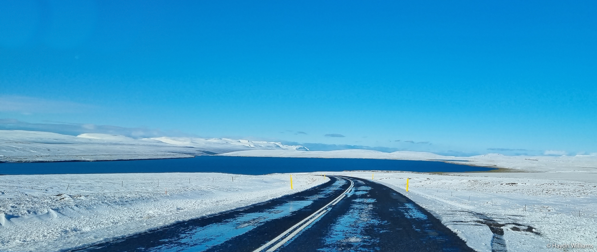 An icy road between snow fields, with a large lake behind. © Haydn Williams 2021