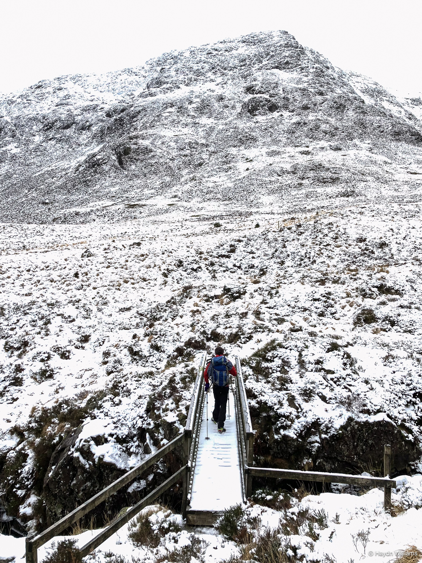 A person crossing a wooden footbridge in the foreground, heading towards a snowy mountain the background. © Haydn Williams 2025