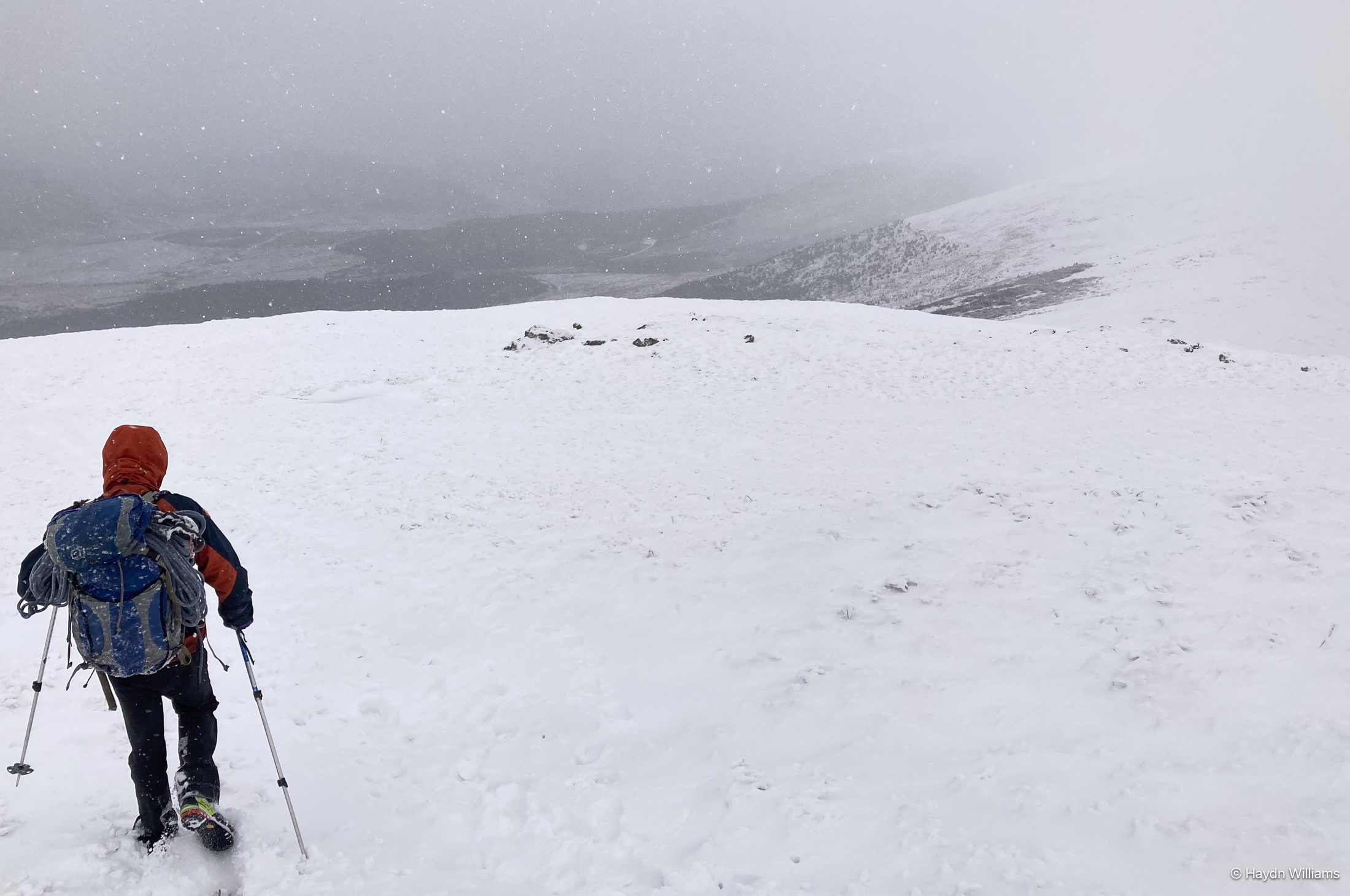 A walker descending a snowy hill as snowflakes fall. © Haydn Williams 2025